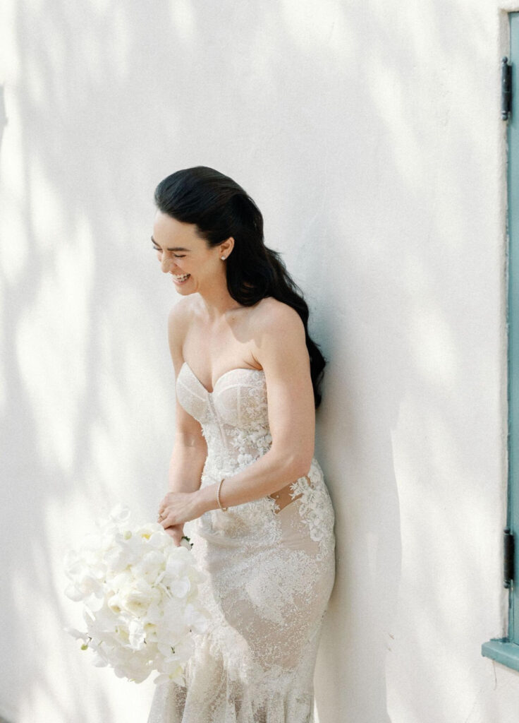A bride in a form-fitting strapless wedding gown and holding a bouquet of white flowers laughs candidly