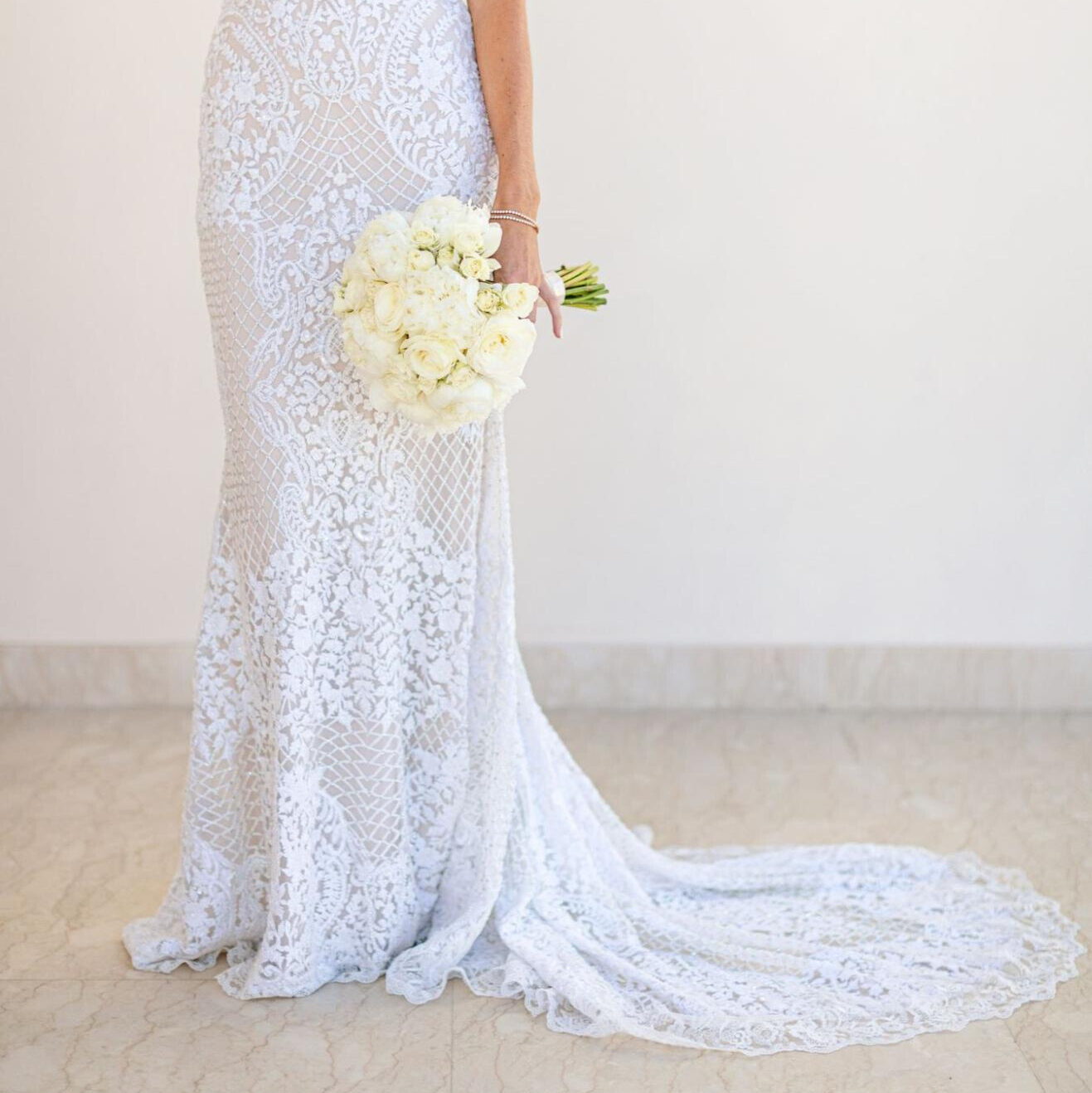 A blonde bride in a fitted lace gown with a train holds a bouquet of white flowers at her side