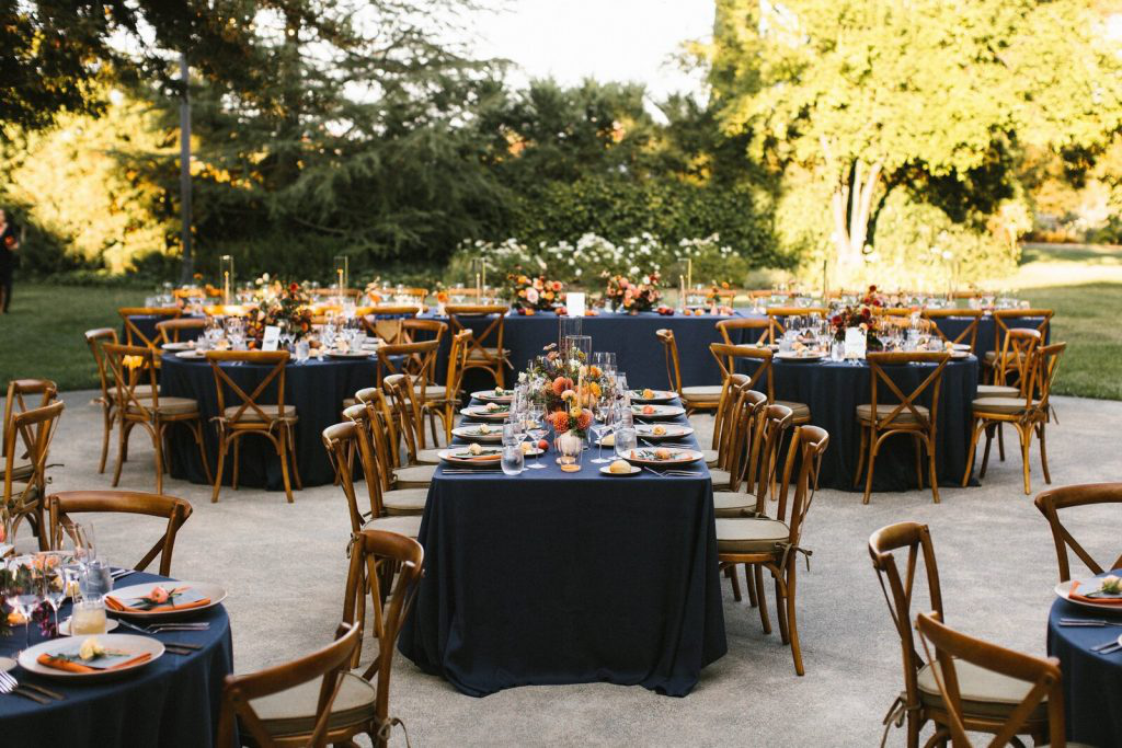 Beautiful outdoor rehearsal dinner setup with tables covered in navy linens, decorated with floral centerpieces, and surrounded by wooden chairs.