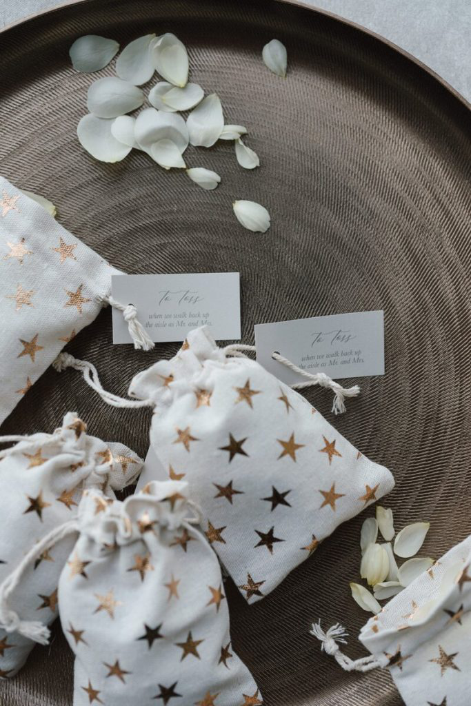 Small white pouches with gold star patterns, filled with wedding favors, displayed on a tray with scattered flower petals.