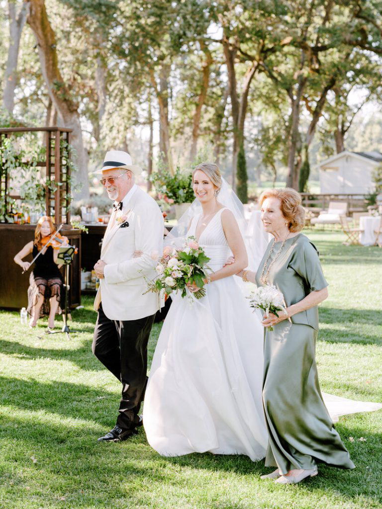 Bride walking with her parents during an outdoor ceremony, surrounded by greenery