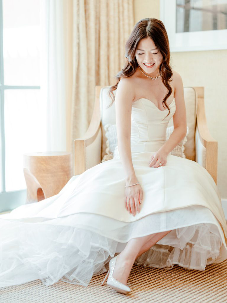 Bride in a strapless satin gown adjusts her elegant shoes, showcasing timeless bridal elegance.