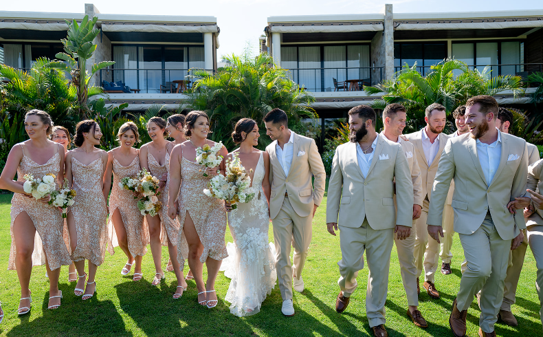 A wedding party including the bride, groom, bridesmaids in champagne dresses, and groomsmen in tan suits walks on green grass outdoors.