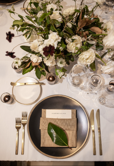 Wedding place setting with neutral linens, gold flatware, and a lush white floral centerpiece