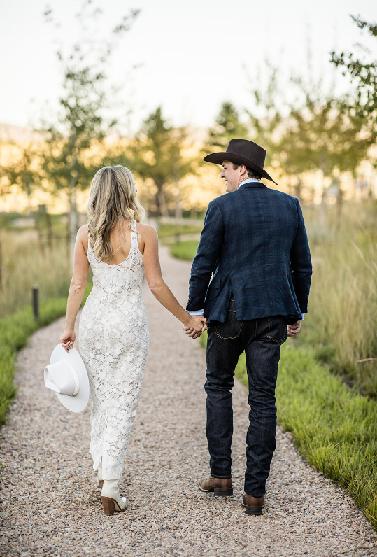Bride in a lace dress and groom in a cowboy hat walking hand in hand on a rustic path in Dallas