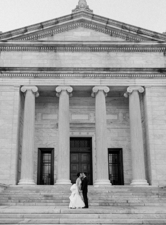 Black and white photo of a bride and groom standing on the grand steps of a neoclassical building with tall columns