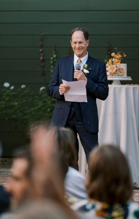 Father of the bride giving a heartfelt wedding speech outdoors with a cake table in the background.