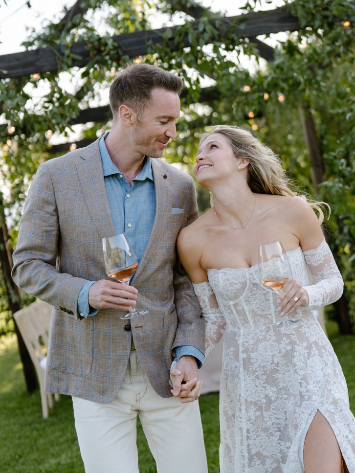 a bride and groom standing outdoors holding a glass of wine.