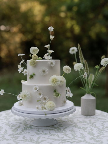 A two-tier wedding cake with white fondant, adorned with small white flowers, green accents, and a contemporary bouquet on a high-end linen outdoor table.