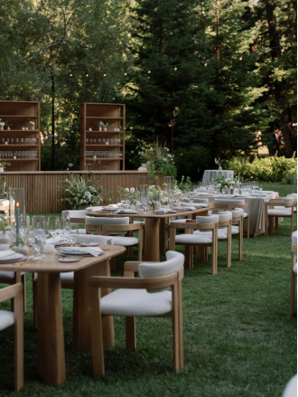 Al fresco wedding dinner setup on a lawn with modern wooden tables, white upholstered chairs, minimalist floral accents, and a custom bar area under tall trees.