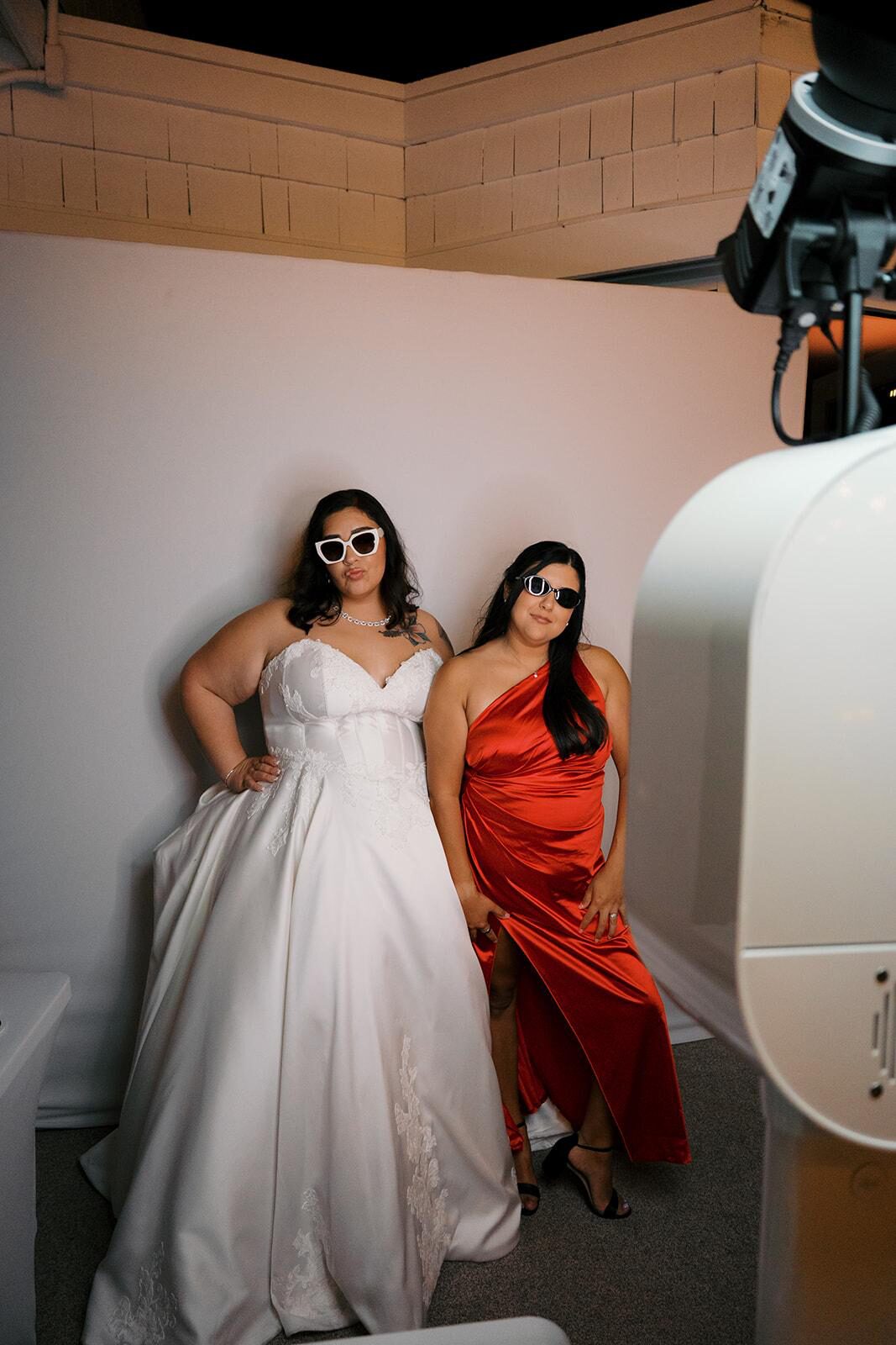 Bride and bridesmaid posing in front of a wedding photobooth, wearing playful glasses