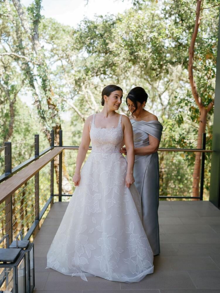 A mother adjusting the white floral wedding gown of her smiling daughter