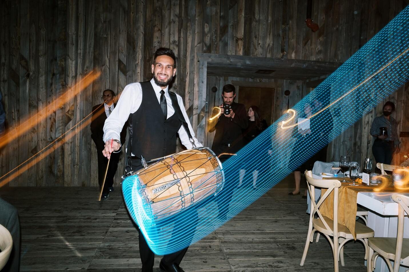 A smiling Dhol player in a vest and tie, drumming energetically
