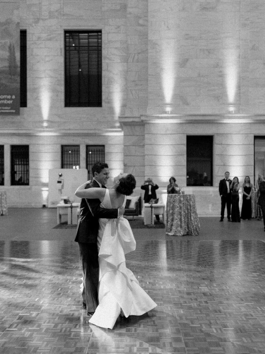 Black and white photograph of the bride and groom during their first dance on a polished wood floor in a large, classic museum hall