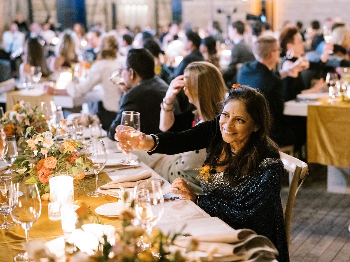 A smiling female wedding guest, wearing a glittering sari, raising a wine glass in a toast at a formal dinner reception table decorated with candles and floral centerpieces