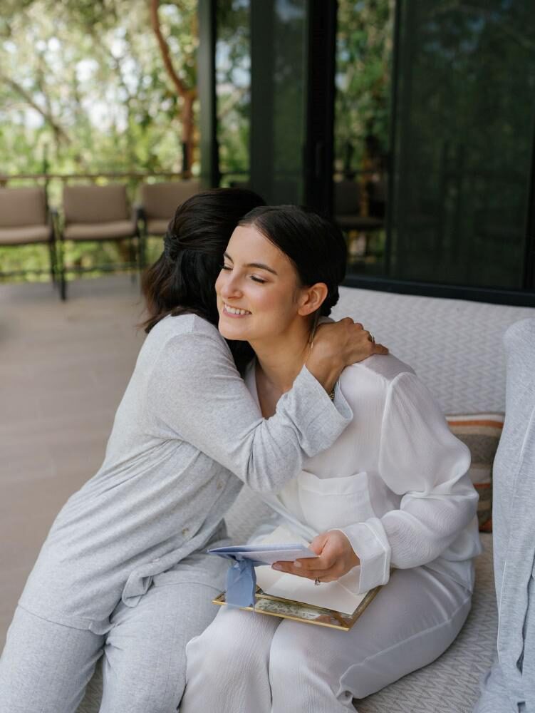 bride in white silk pajamas is hugged by a loved one holding a small photo album or letter on a peaceful outdoor patio.