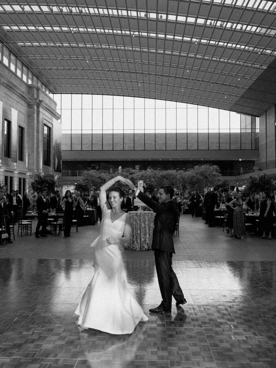 Black and white photo of a bride and groom dancing on a checkered floor in a massive, modern museum atrium