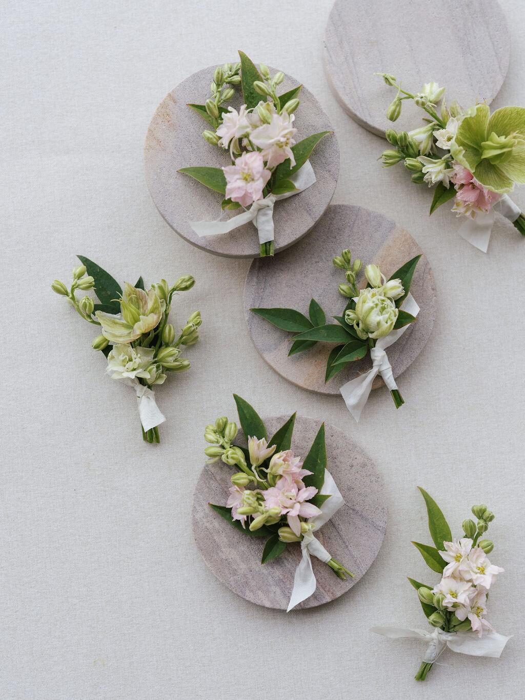 A collection of delicate wedding boutonnieres with pale pink and white flowers and green leaves, each tied with a white ribbon and displayed on round stone plates.
