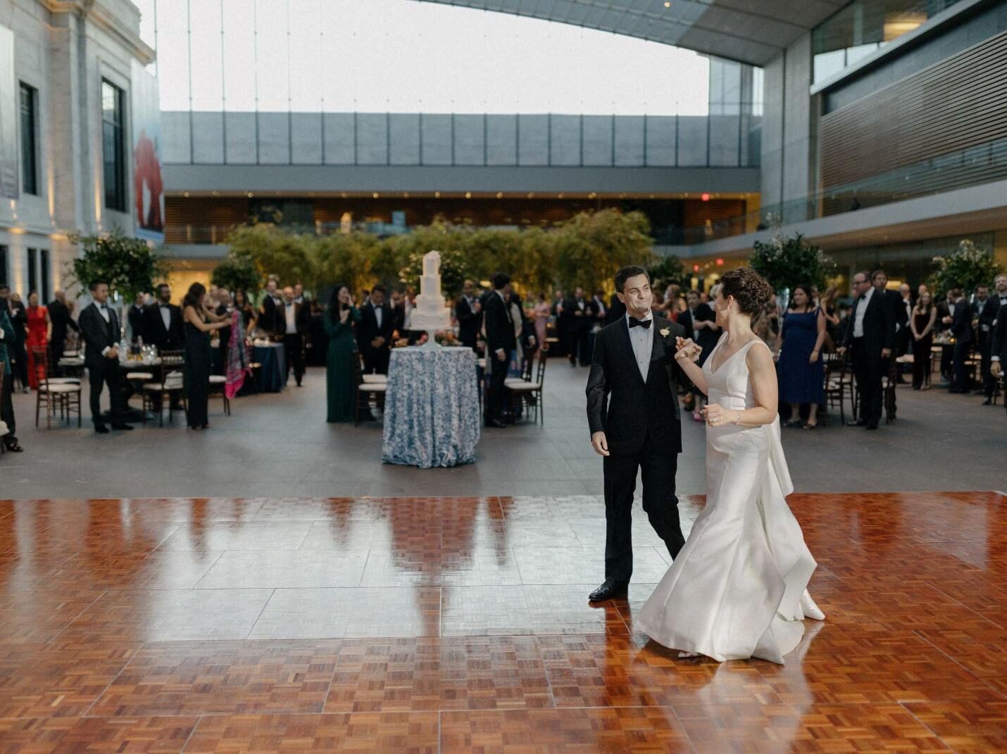 bride and groom standing on a wooden dance floor in a modern museum atrium with a massive glass ceiling. Guests and a multi-tiered wedding cake in the background