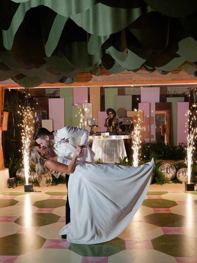 Wedding couple in a dramatic dip kiss on the dance floor with sparkler machines and a DJ in the background.