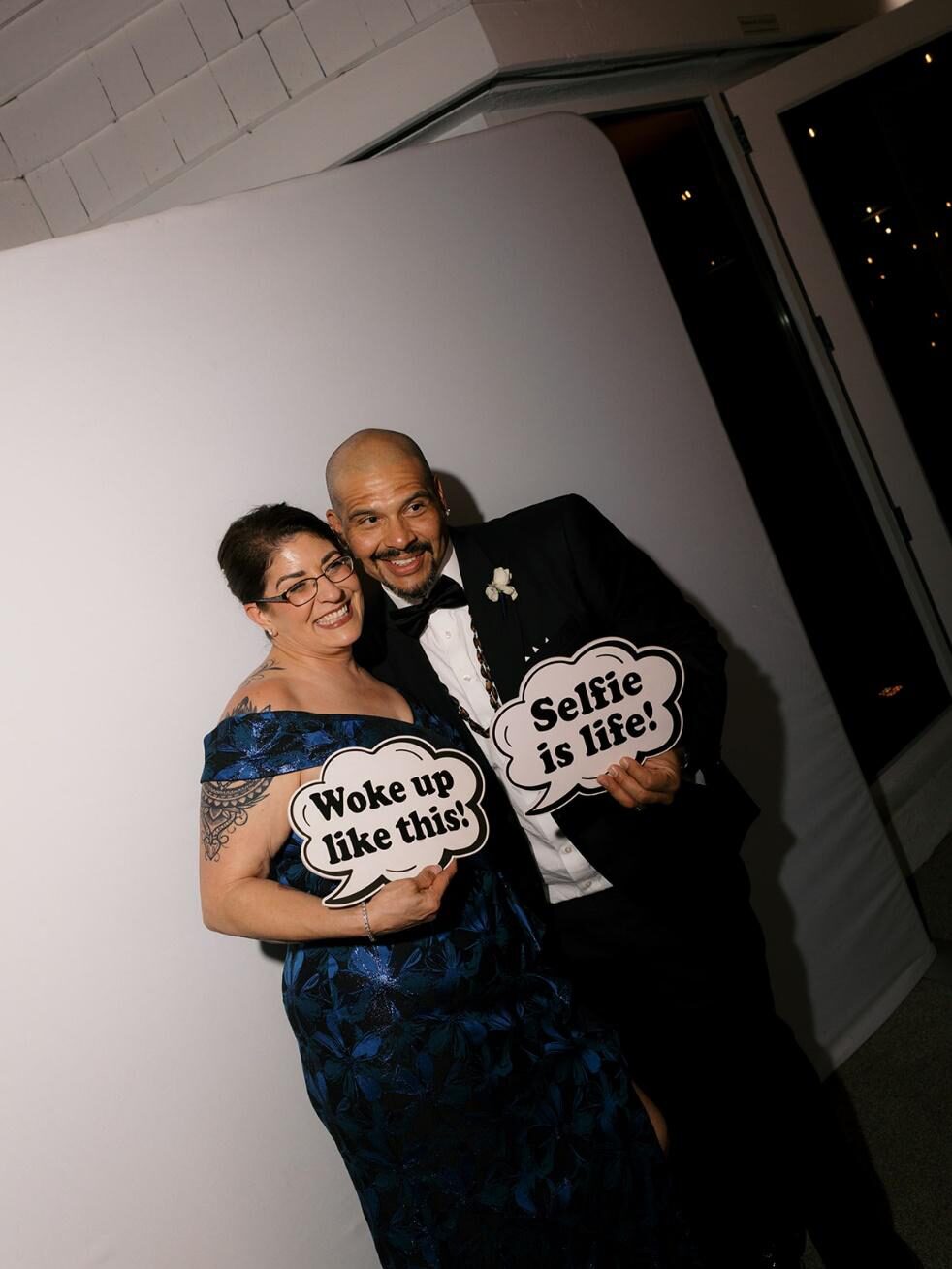 Wedding guests posing with props that read “woke up like this” and “selfie is life” in a photobooth setup.