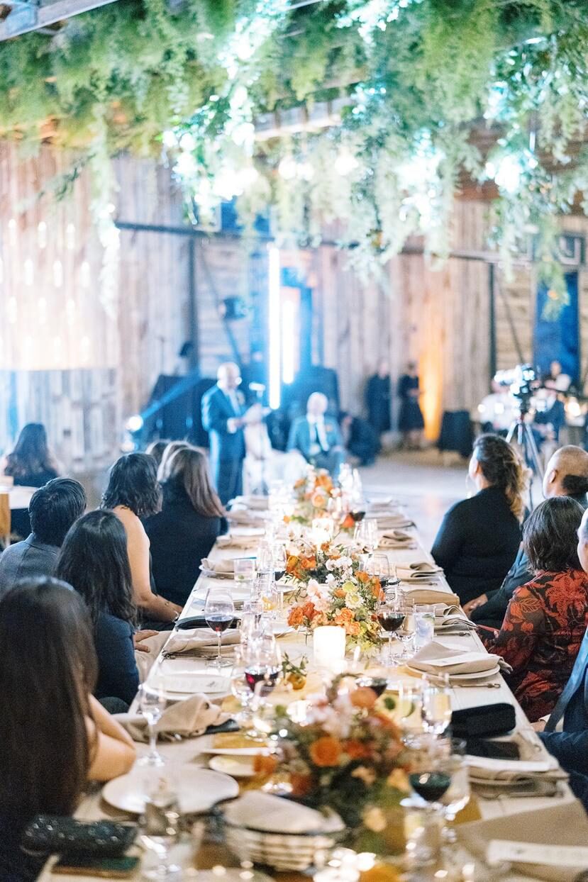 A long wedding reception table beautifully adorned with floral centerpieces in shades of orange and cream, under a lush ceiling installation of greenery and fairy lights.